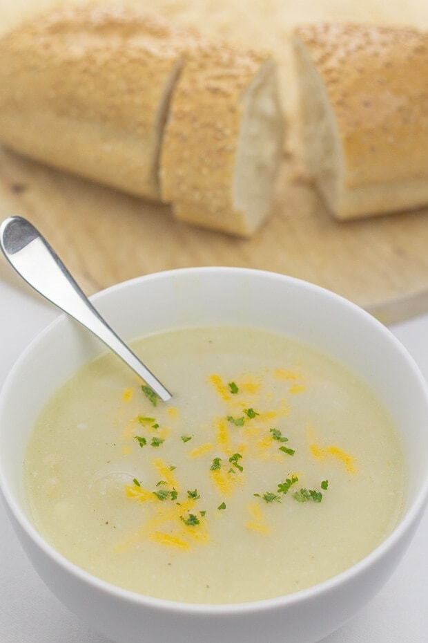 A bowl of cheesy cauliflower and white bean soup with a spoon in. Bread board with a sliced baguette in the background.