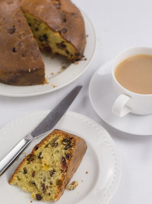 A slice of Dundee cake lying on its side on a plate with a knife. A cup of tea to the right side and the rest of the cake at the top.
