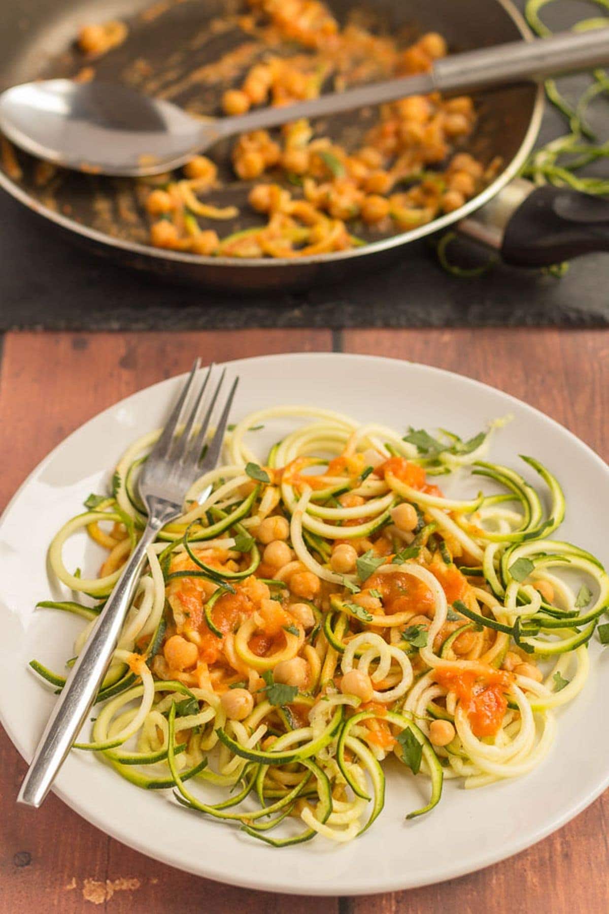 A plate of spiralized courgette spaghetti with chickpeas and the rest of the dish in a pan in the background.