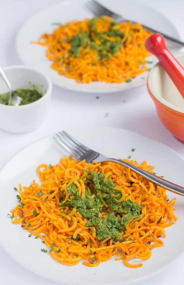 Two plates of baked spiralized sweet potato with spinach pesto with forks on. A dish of pesto and a pestle and mortar in between.