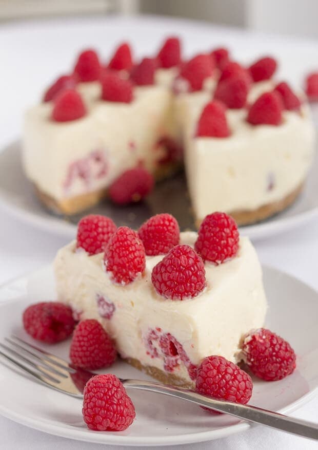 A slice of no bake white chocolate and raspberry cheesecake on a plate with a fork and raspberries to accompany. The rest of the cheesecake in the background.