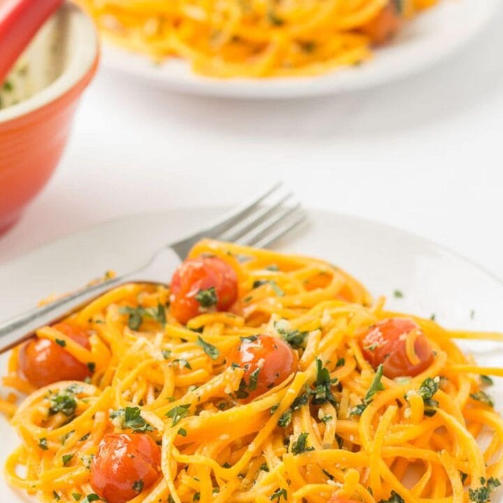 Two plates of butternut squash noodles with spring pesto and roasted tomatoes one in front of the other with forks on. A pestle and mortar in between.