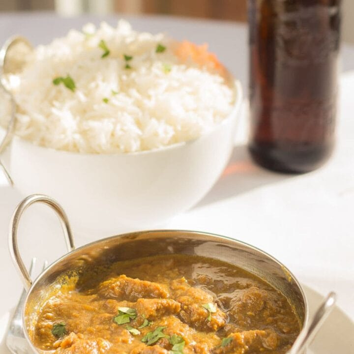 Ceamy coconut beef curry served in a balti dish with a bowl of rice and bottle of beer in the background.