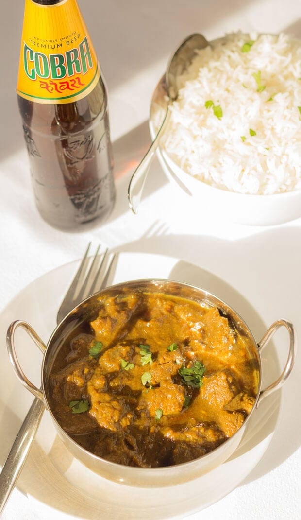 Birds eye view of a balti dish of creamy coconut beef curry on a white plate with a fork beside it. A bottle of beer and bowl of rice with a spoon in the background.
