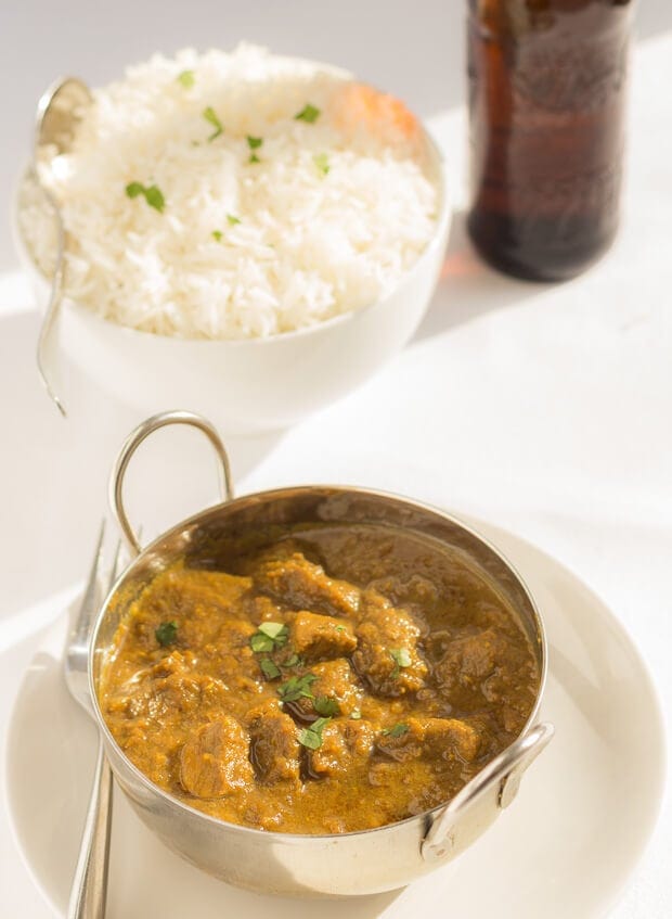Close up of a balti dish of creamy coconut beef curry with a bowl of rice and bottle of beer in the background.