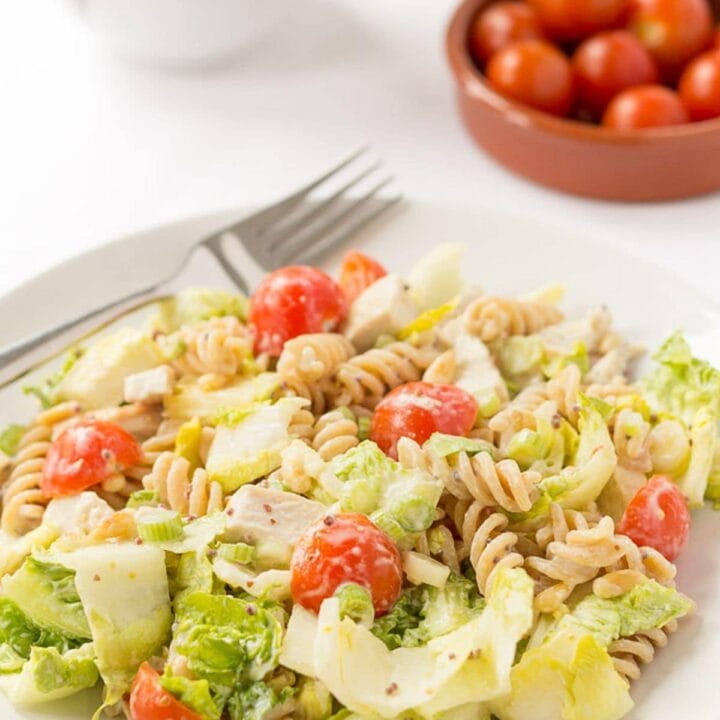 A plate of honey mustard turkey pasta salad with knife and fork on. A dish of cherry tomatoes and white jug in the background.