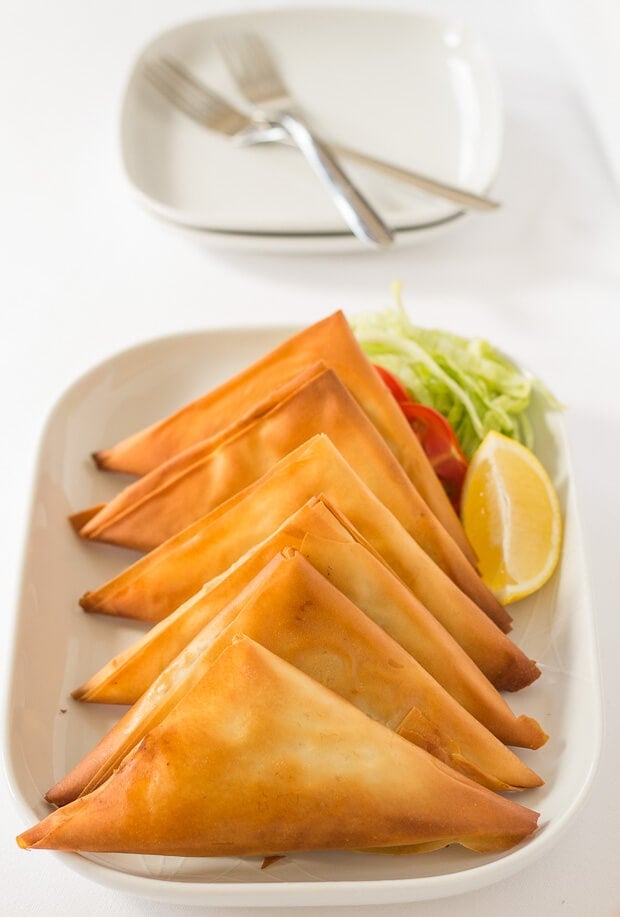 Close up of a serving dish with six turkey filo pastry samosas on. Garnished with salad and a slice of lemon. Two plates and forks in the background. are nearly a third of the calories of those you might find in a restaurant. They’re filled with a delicious lightly spiced turkey mince, then baked rather than deep fried, making them a great quick healthy meal!
