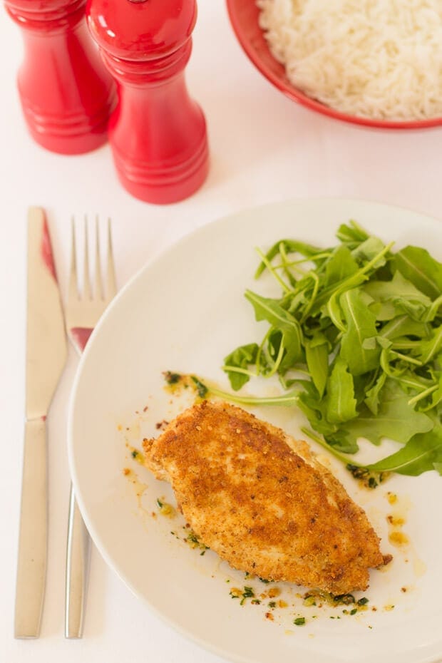 Birds eye view of easy baked chicken kiev served on a plate with rocket salad. Knife and fork beside. Salt and pepper cellars and a bowl of rice at the top.