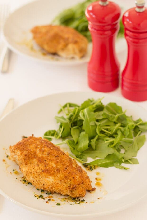 Close up of a plate of easy baked chicken kiev served with rocket. Salt and pepper cellars and another plate with a kiev and rocket on in the background.