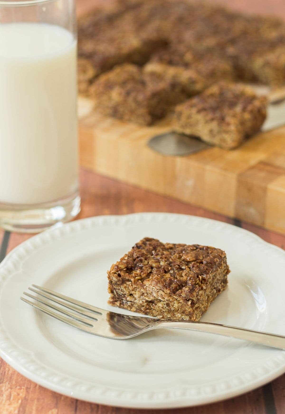 A plate with a no bake chocolate oatmeal bar on. Fork to the side and a glass of milk and chopping board with the rest of the oat bars on in the background.