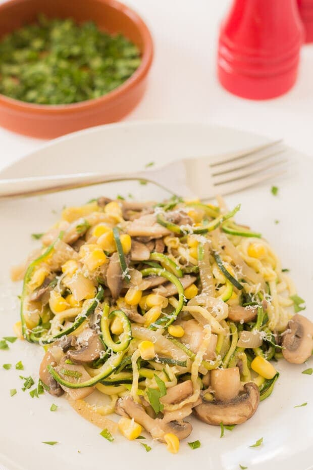 Close up of a plate of zucchini mushroom pasta with a fork on. A dish of chopped coriander and salt and pepper cellars in the background.