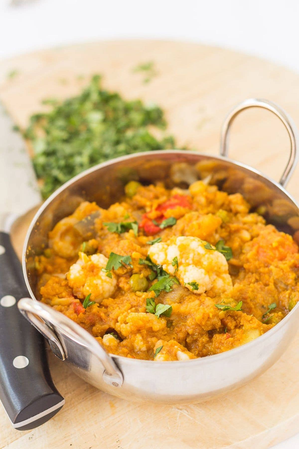 Cauliflower dhansak served in a balti dish placed on a chopping board with a knife and chopped coriander alongside.