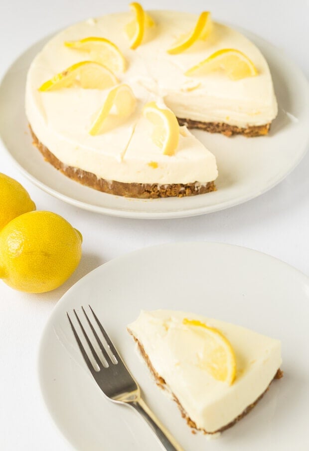 A slice of lemon crunch cheesecake on a plate with a fork to the side. The rest of the lemon cheesecake at the top and two lemons in between.