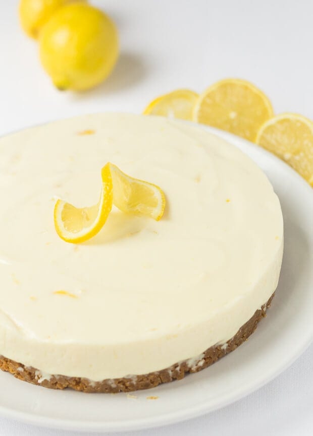 A whole uncut lemon crunch cheesecake with a twisted slice of lemon on top and sliced lemons in the background as decoration.