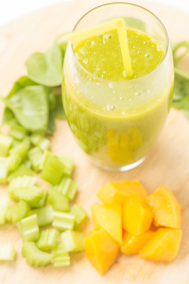 Birds eye view of an orange mango green smoothie on a chopping board with chopped mango, celery and spinach arranged around.