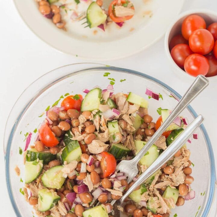 Birds eye view of a salad bowl of borlotti bean and tuna salad with serving utensils in at the bottom and a plate with a serving of the salad and a fork on at the top.