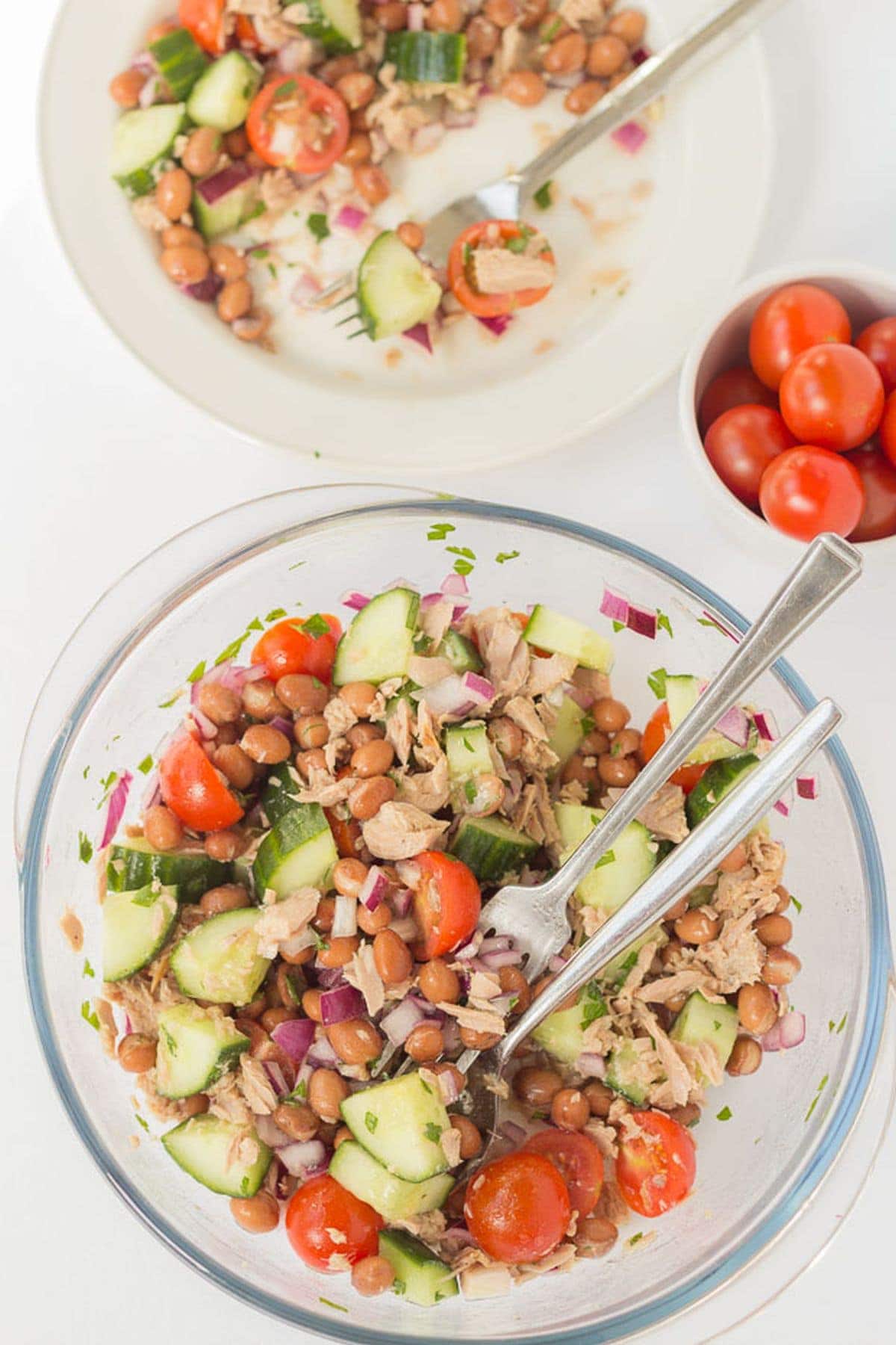 Birds eye view of a salad bowl of borlotti bean and tuna salad with serving utensils in at the bottom and a plate with a serving of the salad and a fork on at the top.