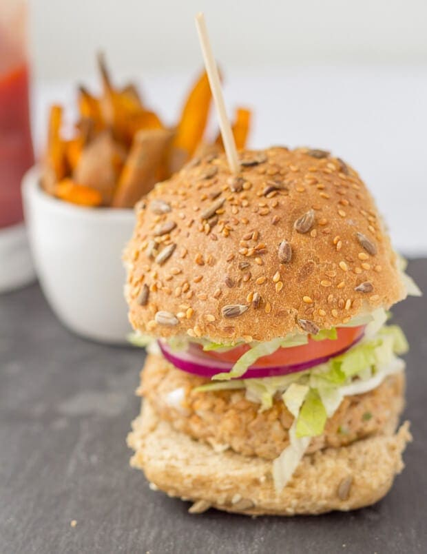 Close up of a tuna burger on a bun garnished with iceberg lettuce, sliced tomato and red onion. Bowl of sweet potato fries in the background.