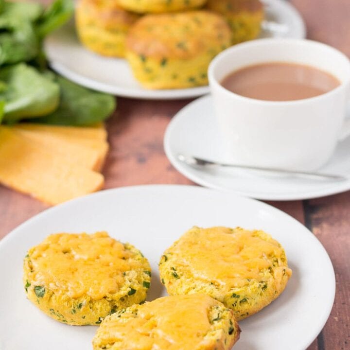 A plate of sweet potato spinach scones cut in half with melted cheese on. A cup of tea and the rest of the whole spinach scones on another plate in the background.