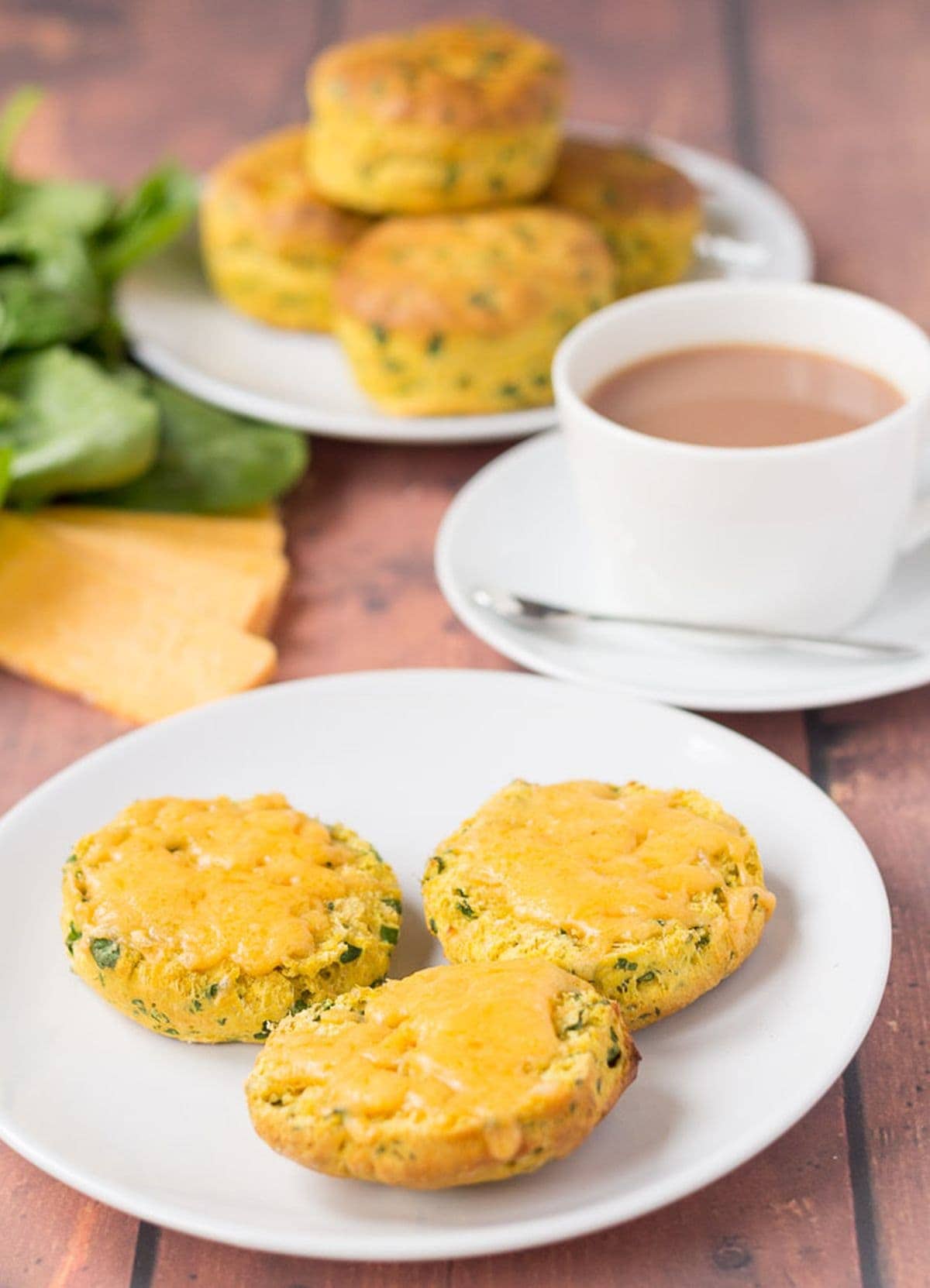 A plate of sweet potato spinach scones cut in half with melted cheese on. A cup of tea and the rest of the whole spinach scones on another plate in the background.