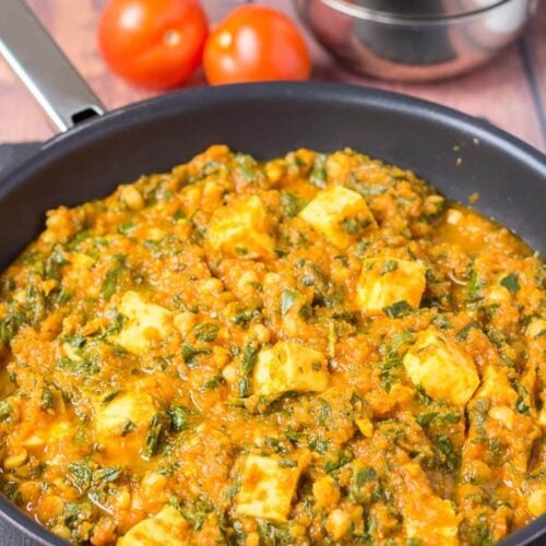 A pan of cooked paneer and chickpea curry with two whole tomatoes and two stacked balti dishes in the background for decoration.