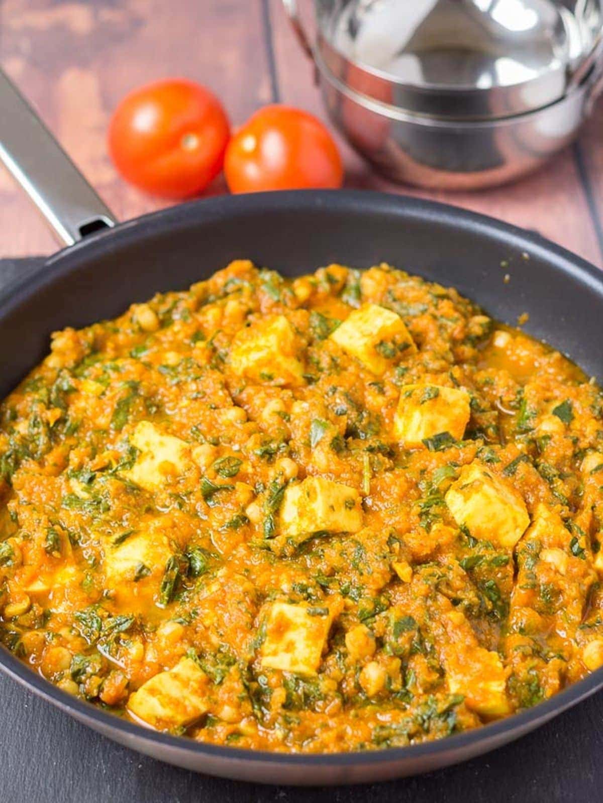 A pan of cooked paneer and chickpea curry with two whole tomatoes and two stacked balti dishes in the background for decoration.