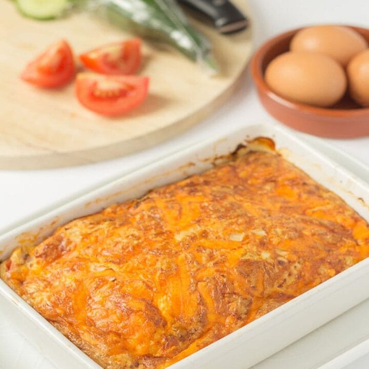 A casserole dish of cooked baked cheese and tomato omelette just removed from oven. Chopping board with salad on and dish of eggs in the background.
