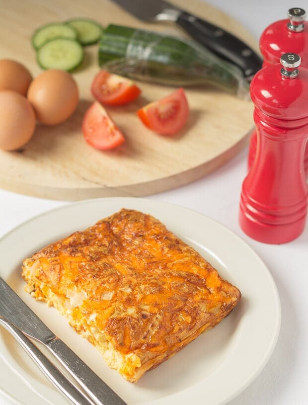 A portion of baked cheese and tomato omelette served on a plate with a knife and fork beside. Chopping board with eggs, sliced cucumber and quartered tomatoes in the background. Salt and pepper cellars to the side.