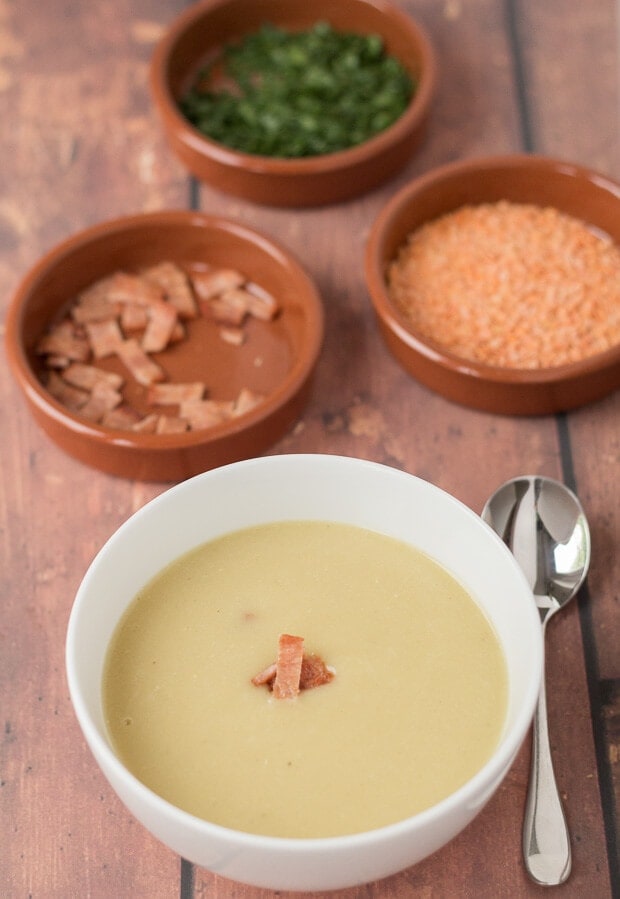 A bowl of lentil and potato soup topped with crispy bacon. Soup spoon to the side and dishes of lentils, parsley and chopped bacon in the background.