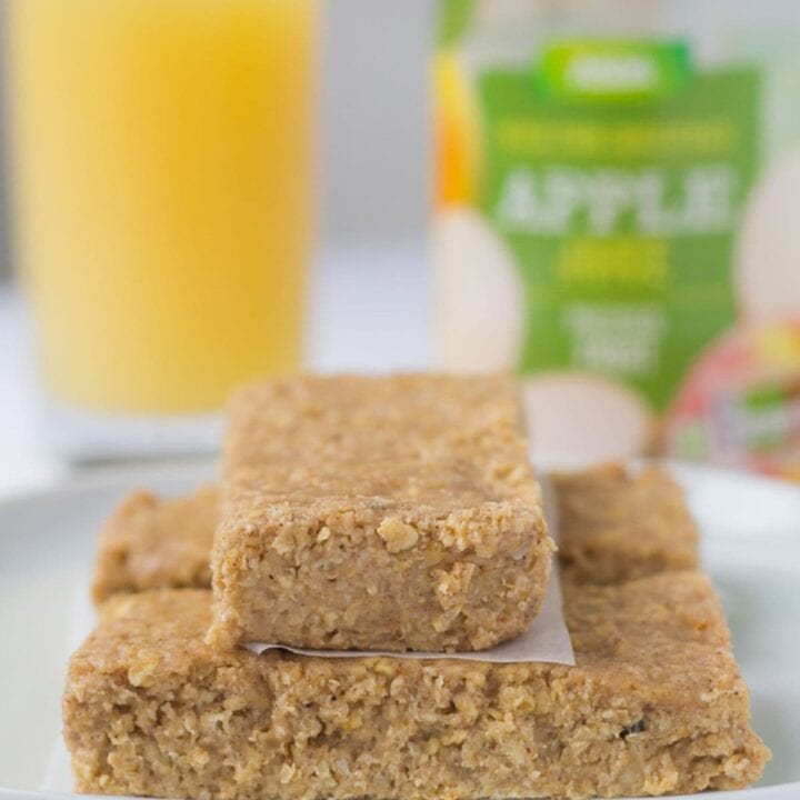 A plate of three no bake apple and cinnamon oat bars with a glass of apple juice and a plastic bottle juice bottle in the background.