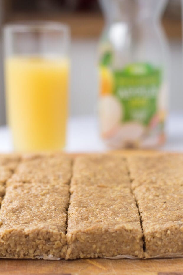 Close up of a bread board of no bake apple and cinnamon oat bars. Glass of apple juice and plastic bottle in the background.