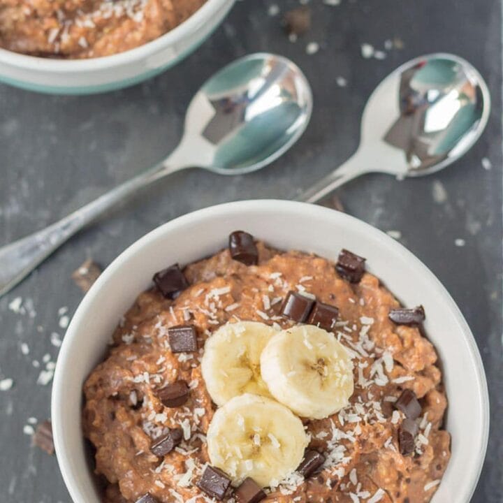 Birds eye view of a bowl of chocolate chip banana coconut porridge on a black slate with two spoons beside.