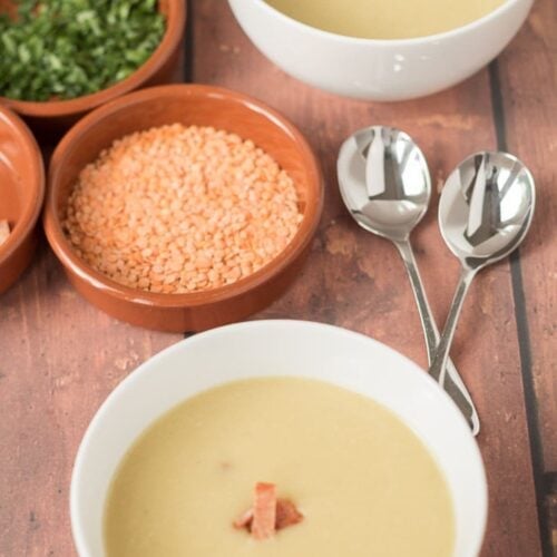 Two bowls of lentil and potato soup with dishes of lentils and herbs between. The bowls of soup topped with crispy bacon.