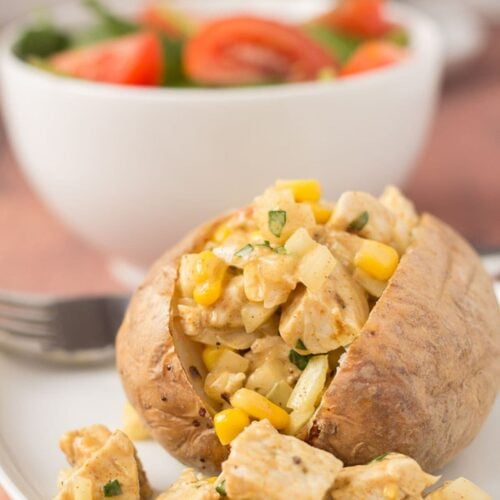 A baked potato with Coronation turkey on a plate with the filling falling out. A bowl of salad in the background.
