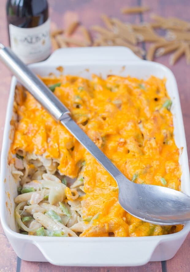 A casserole dish of cheesy bacon pasta bake with green beans with a portion removed and the serving spoon on top. A bottle of wine and some dried pasta scattered about in the background.