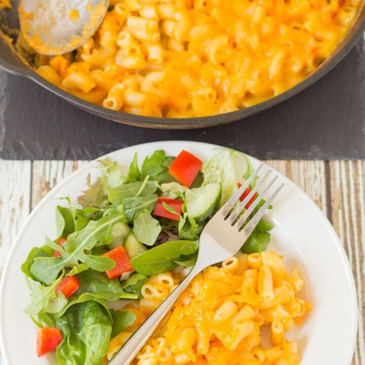 Birds eye view of a plate with skilled baked macaroni and cheese on with a side salad at the bottom. Skillet with serving spoon in at the top.