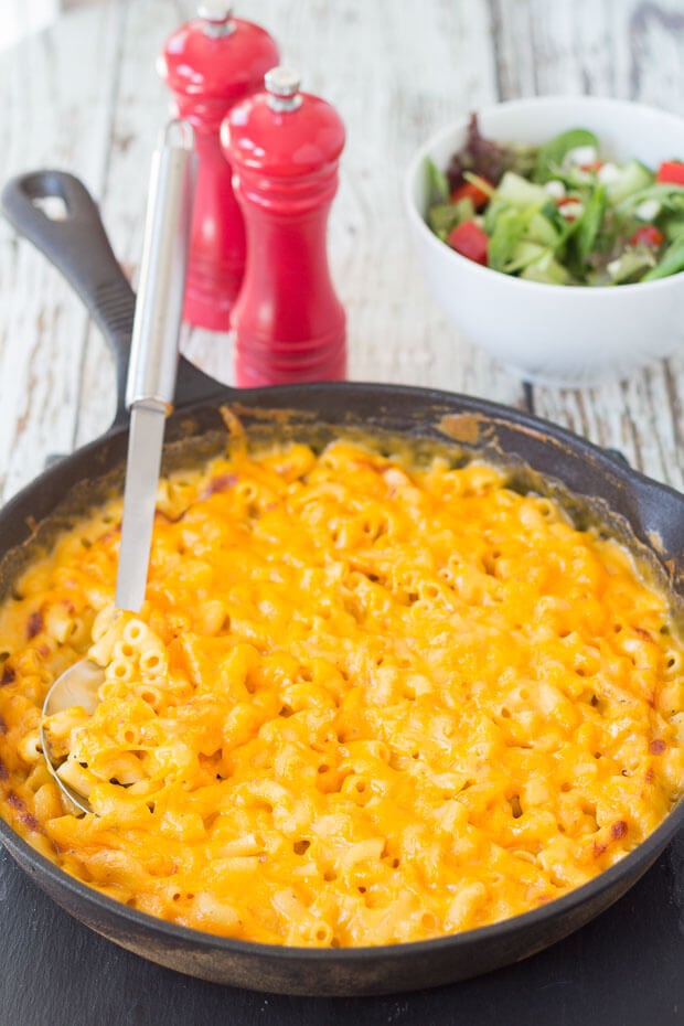 Skillet baked macaroni and cheese with a serving spoon in. Salt and pepper cellars and a bowl of salad in the background.
