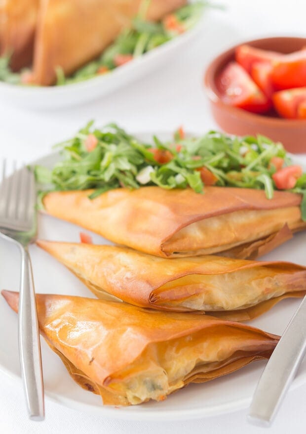 Threee tomato basil mozzarella filo parcels on a plate with a knife and fork and some lettuce and finely chopped tomatoes. A dish of tomatoes and another plate in the background.
