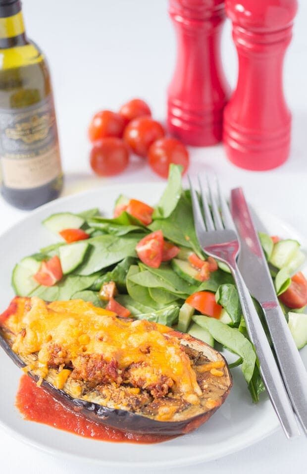 A plate of half a baked aubergine with red pesto and cheese and salad and a knife and fork on. A bottle of wine, cherry tomatoes and salt and pepper cellars in the background.