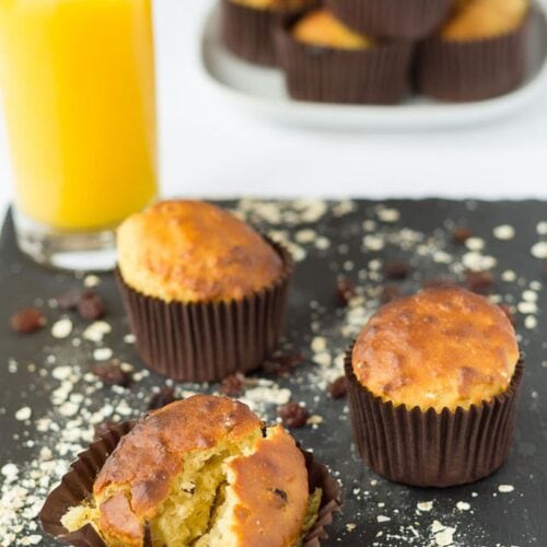 Three raisin oat breakfast muffins on a black slate. Glass of orange juice and a stack of the muffins on a plate in the background.