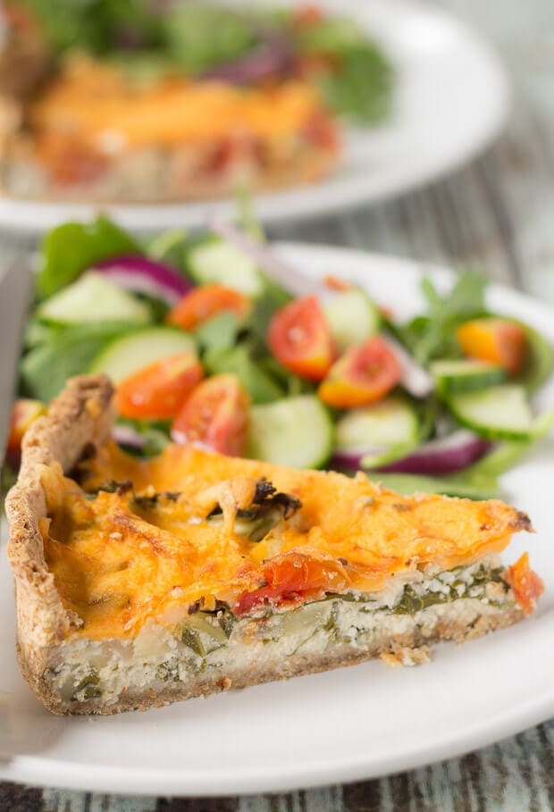 A slice of cheesy tomato and kale quiche on a plate with a side salad. Another duplicate plate in the background.