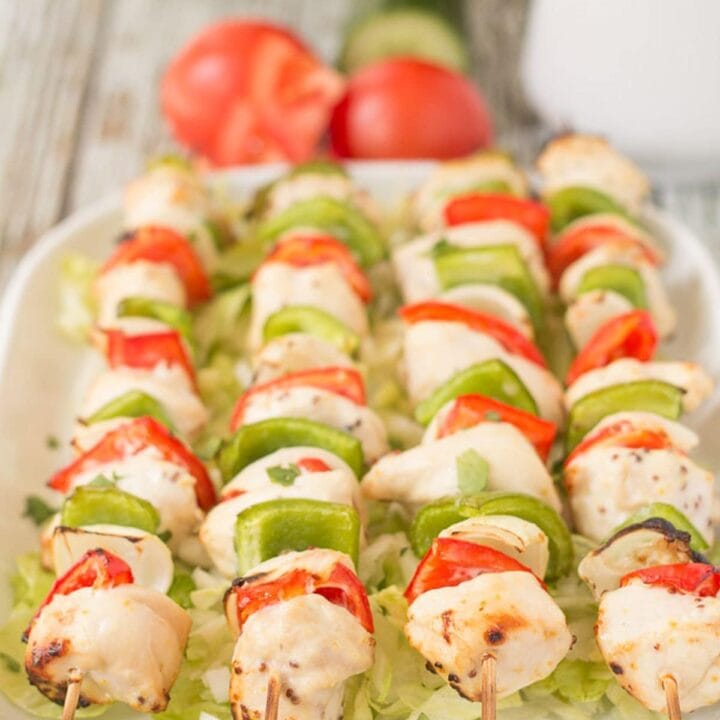 Four honey mustard chicken skewers on a serving platter with two tomatoes, a cucumber and a jug in the background.