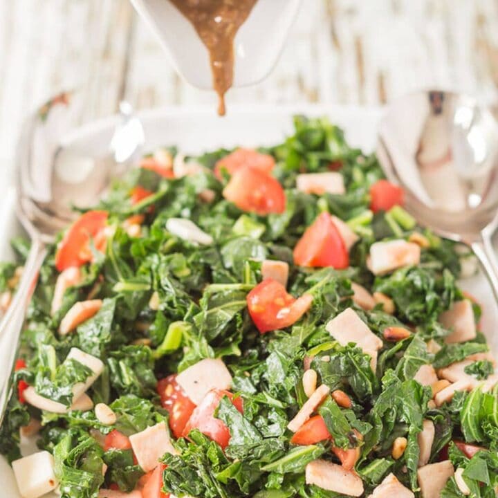 Balsamic dressing being poured from a jug onto a platter of kale and turkey bacon salad.