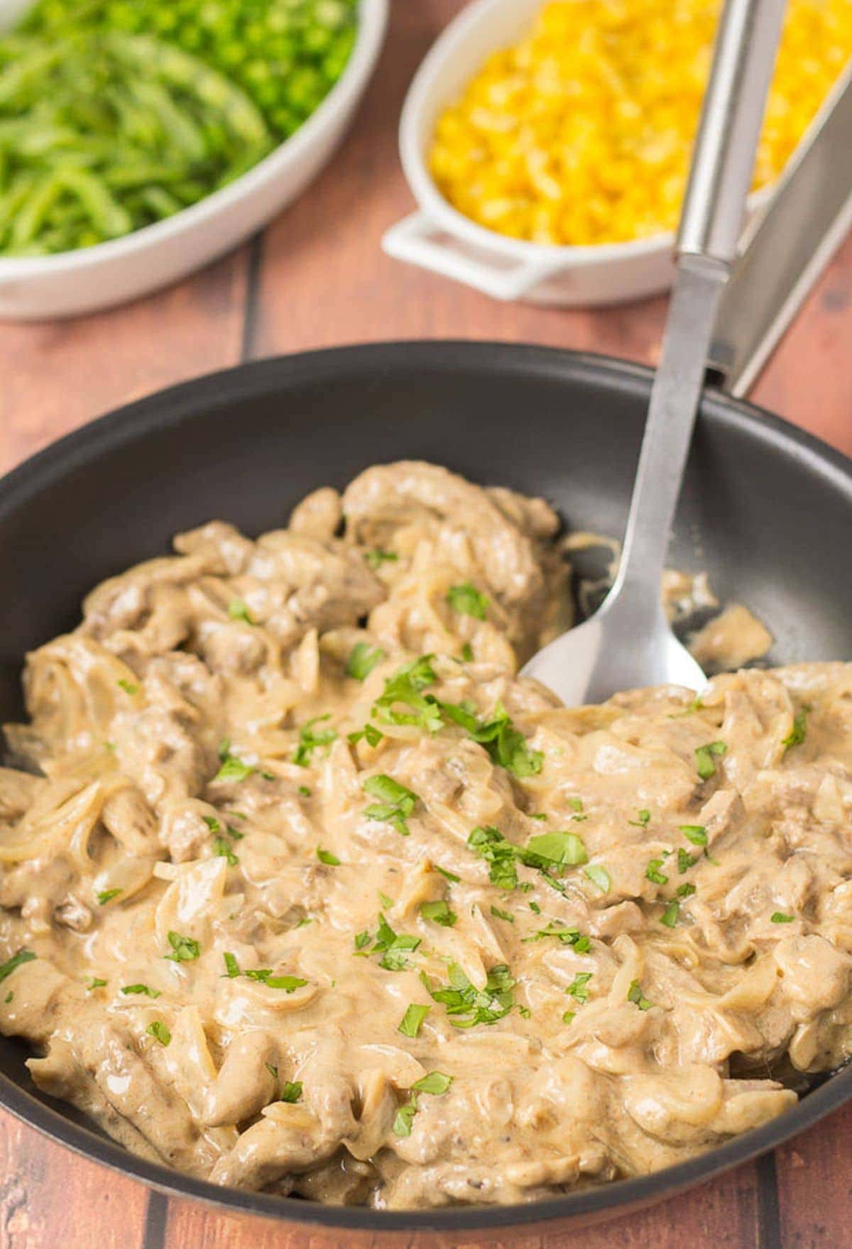 A pan of cooked quick healthy beef stroganoff with a serving spoon in. Side dishes of green beans and sweetcorn in the background.
