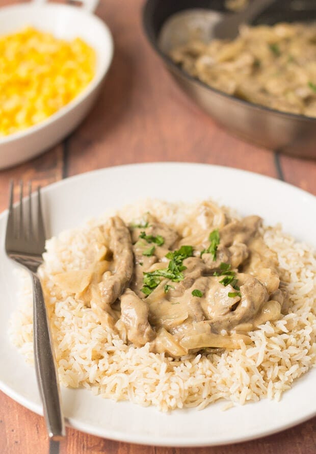 Close up of a plate of quick healthy beef stroganoff served on a bed of wholegrain rice. Fork to the side. Dish of sweetcorn and cooking pan in the background.