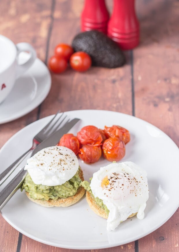 A plate of smashed avocado and poached eggs on muffins. Served with cherry tomatoes and a knife and fork to the side. In the background a coffee cup, salt and pepper cellars and half an avocado.