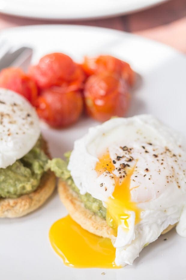 Close up of a plates of smashed avocado and poached eggs on muffins with cherry tomatoes and a knife and fork on. One of the eggs with a burst yolk.