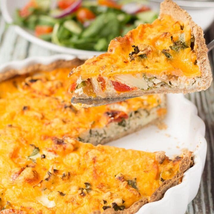 A slice of cheesy tomato and kale quiche being lifed from the quiche dish. A plate with salad on in the background.