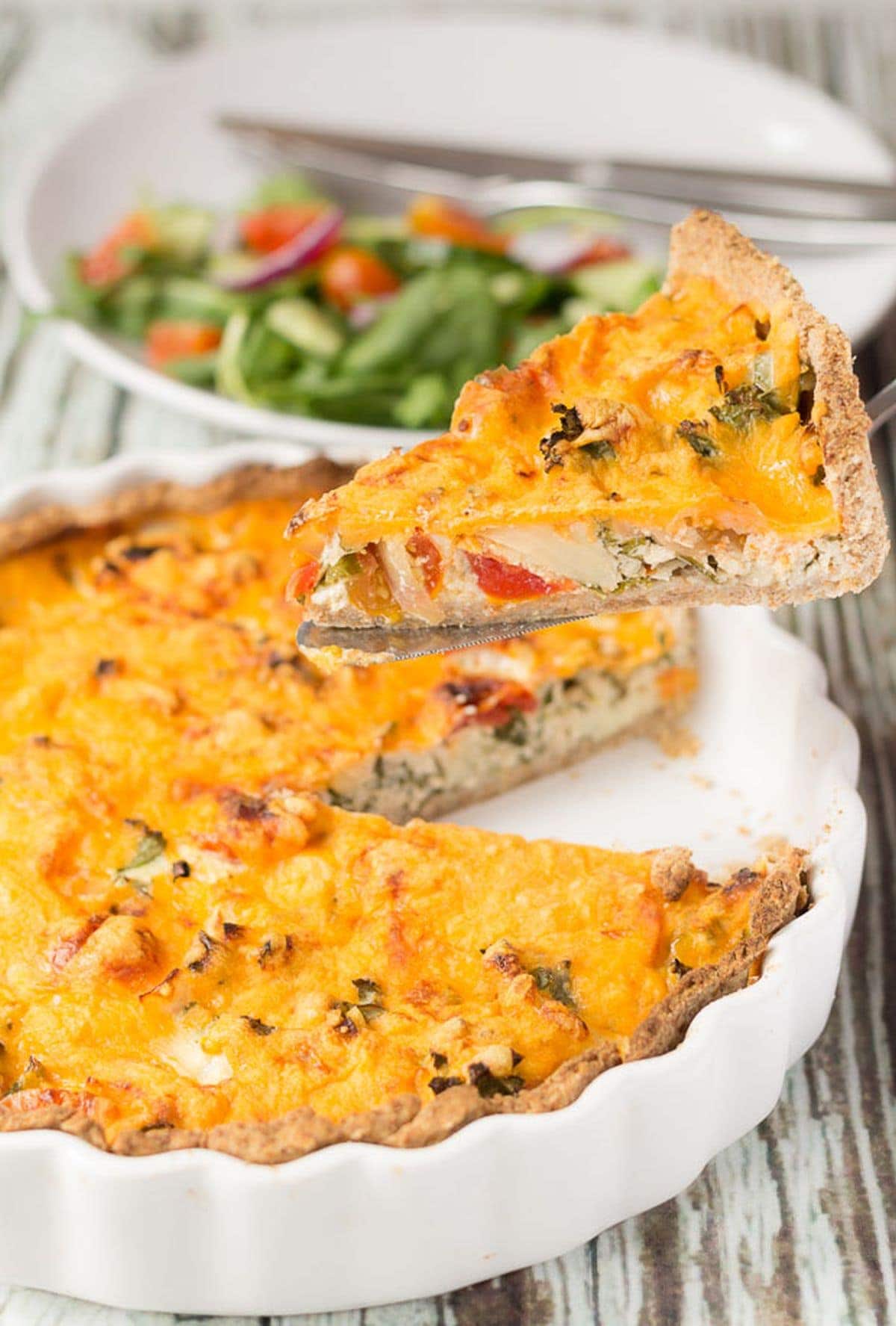 A slice of cheesy tomato and kale quiche being lifed from the quiche dish. A plate with salad on in the background.