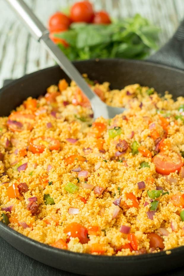A pan of Mediterranean chorizo couscous with a serving spoon in. Basil and cherry tomatoes in the background as decoration.